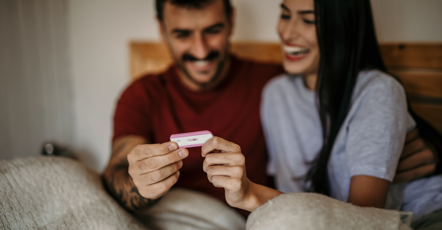A joyful couple shares a moment of excitement while looking at a pregnancy test result in bed.