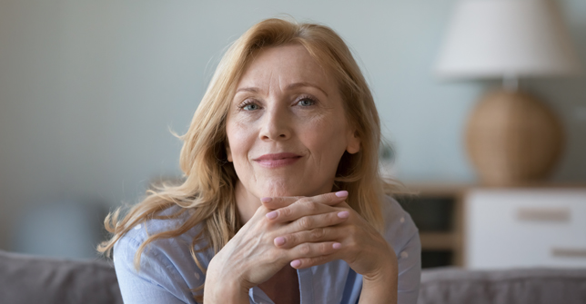 Smiling middle-aged woman with long hair resting her chin on her hands, conveying a sense of calm and reflection.