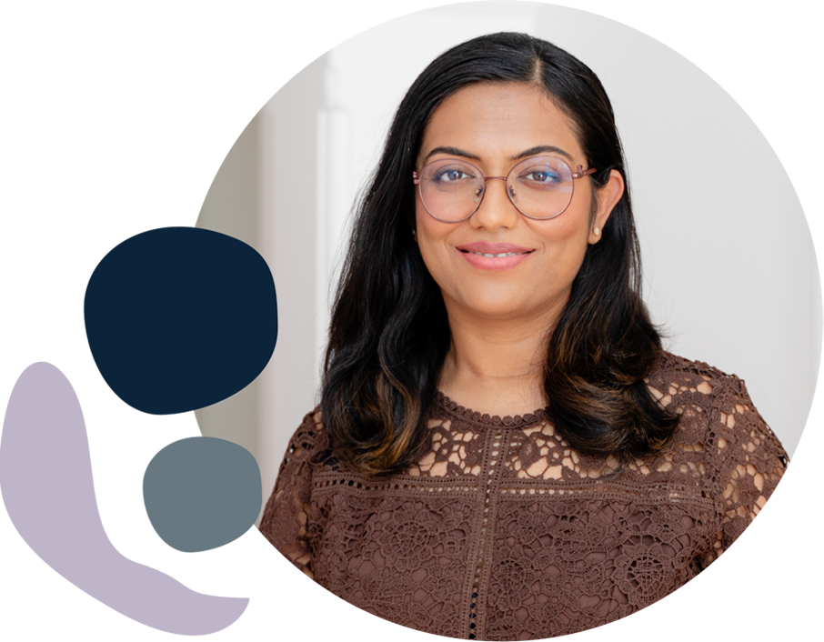 A woman with long dark hair and round glasses wears a brown lace top, posing against a light background with abstract dark blue and grey shapes.